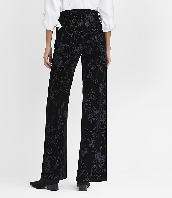 A person wearing a white blouse and black floral-patterned trousers stands against a plain white background.