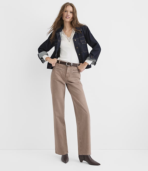 A woman wearing a dark denim jacket, white lace-trim top, and brown trousers stands against a plain white background.