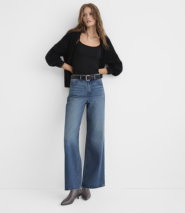 A woman wearing a black cardigan, black top, and wide-leg blue jeans stands against a plain white background.