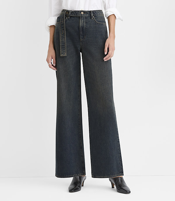 A person wearing a white shirt, wide-leg dark blue jeans with a belt, and black pointed heels stands against a plain white background.