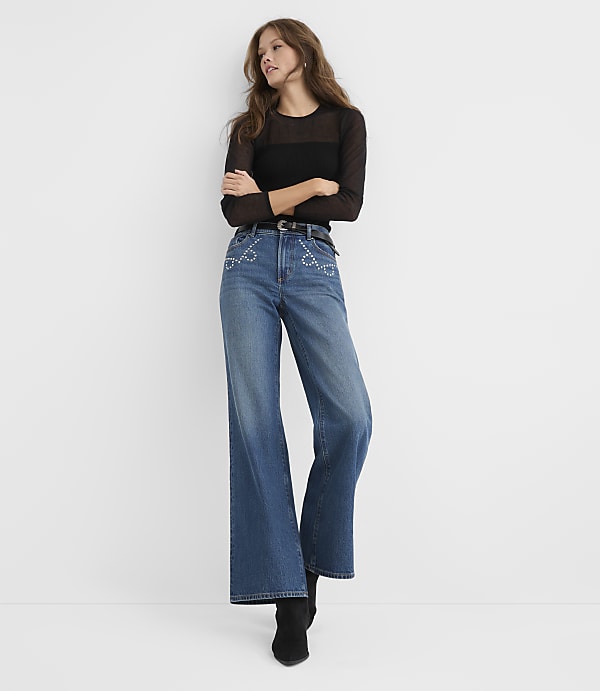 A woman wearing a sheer black top and wide-leg blue jeans stands against a plain white background.