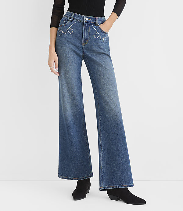 A person wearing a black top, wide-leg blue jeans with rhinestone details, and black boots stands against a plain white background.