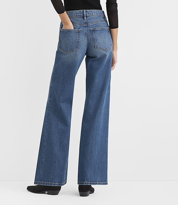 A person wearing a black top, wide-leg blue jeans, and black shoes stands against a plain white background.