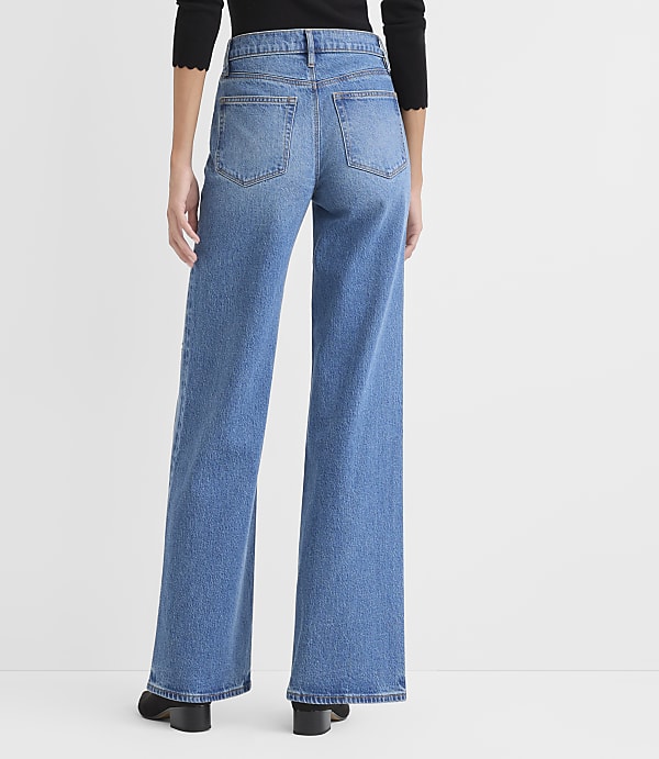 A person wearing a black top and wide-leg blue jeans stands against a plain white background.