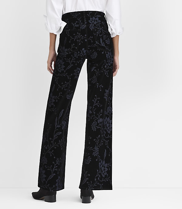 A person stands with their back to the camera wearing a white blouse and floral-patterned black velvet trousers against a plain white background.