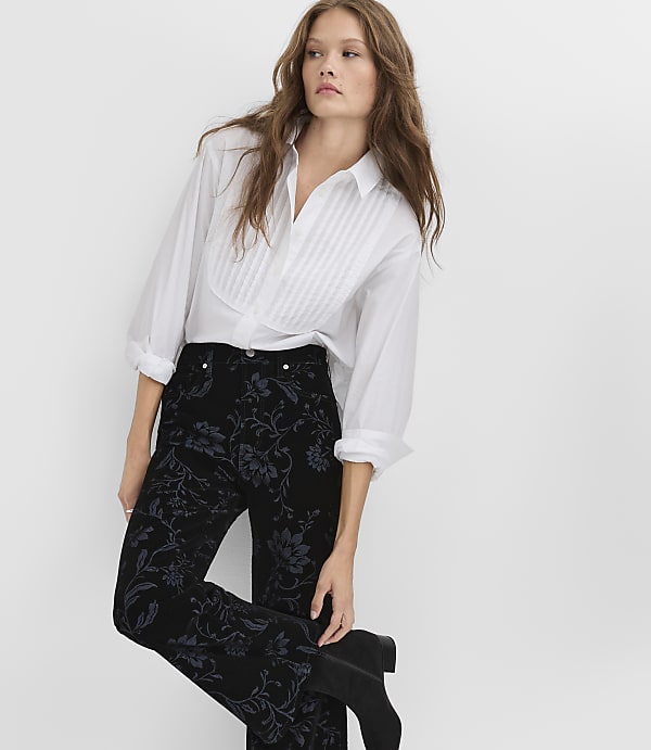 A woman wearing a white pleated shirt and black floral trousers stands against a plain white background.