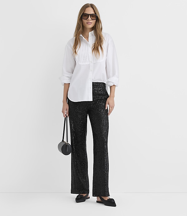 A woman wearing a white button-down shirt, shimmering black trousers, and sunglasses stands against a plain white background.