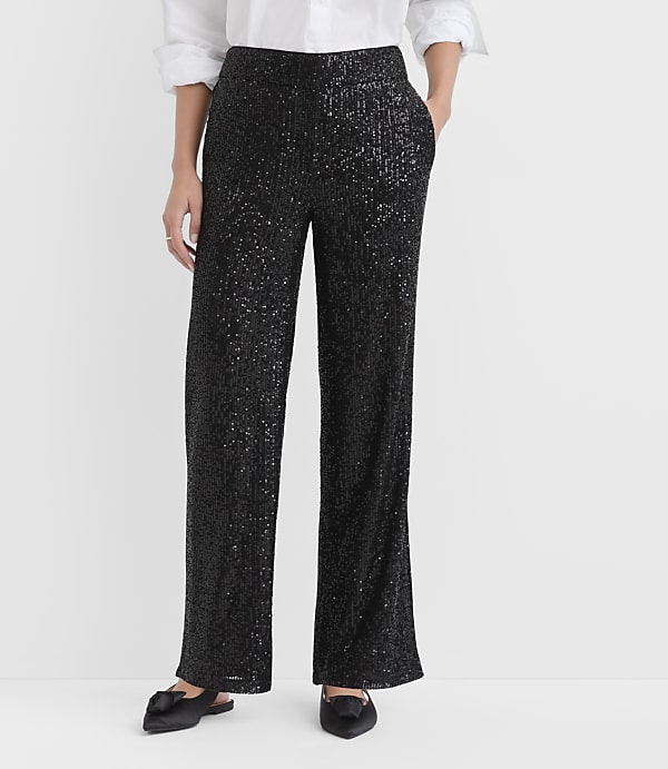 A person wearing a white shirt, shimmering black sequined trousers, and black pointed flats stands against a plain white background.