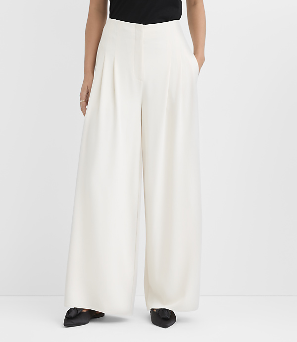 A person wearing a black top and wide-leg white trousers stands against a plain white background.