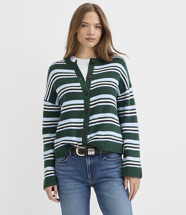 A woman wearing a green and blue striped cardigan and blue jeans stands against a plain white background.