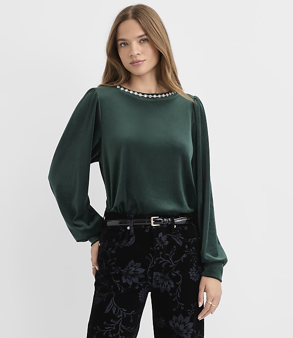A woman wearing a dark green blouse and black floral trousers stands against a plain white background.