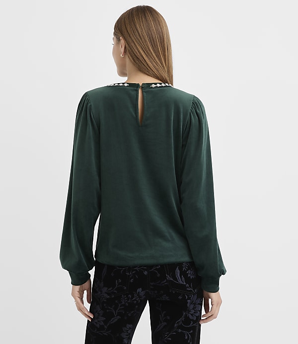 A woman wearing a dark green velvet top and black floral pants stands with her back to the camera against a plain white background.