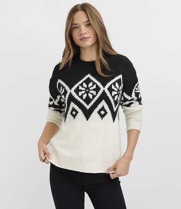 A woman wearing a black and white patterned sweater and black pants stands against a plain white background.