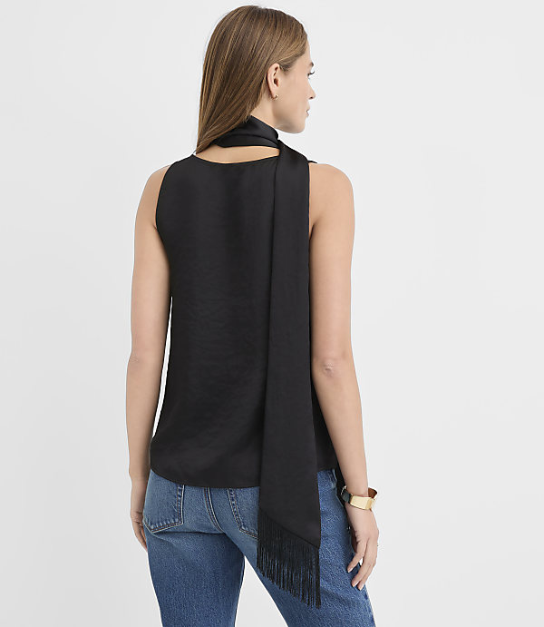 A woman wearing a black sleeveless top with a matching scarf and blue jeans stands against a plain white background.