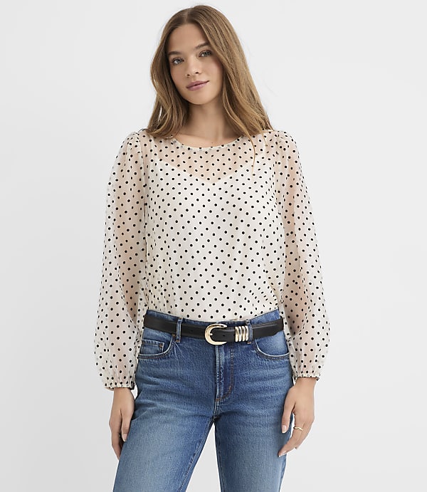 A woman wearing a white polka-dot blouse and blue jeans stands against a plain white background.