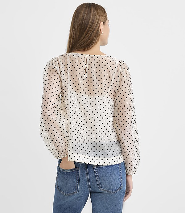 A woman wearing a white polka-dot blouse and blue jeans stands with her back to the camera against a plain white background.