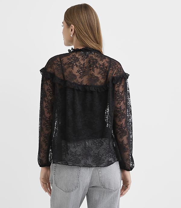 A woman stands with her back to the camera wearing a black lace blouse and grey jeans against a plain white background.