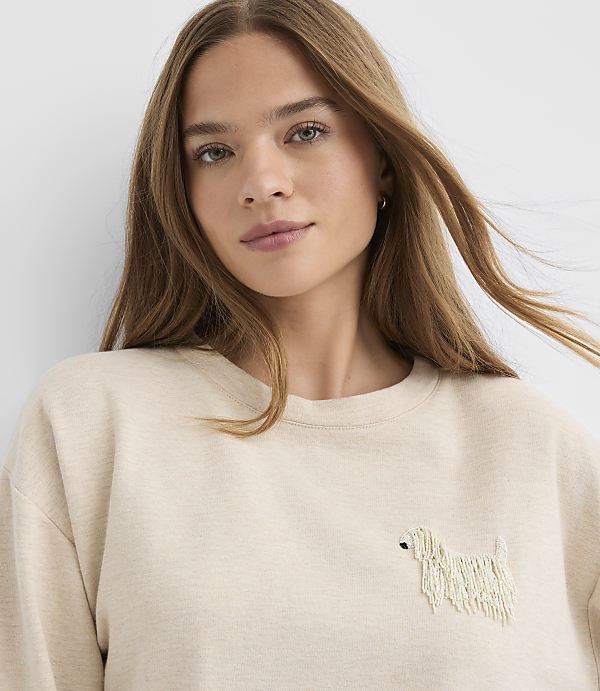 A woman with long brown hair wearing a beige sweatshirt with a small dog embroidery stands against a plain white background.
