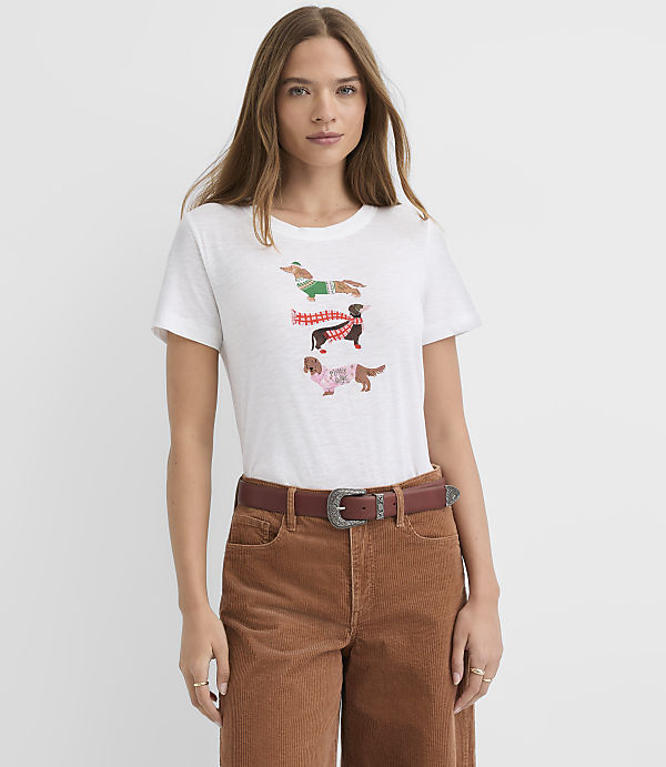 A woman wearing a white t-shirt with dachshund graphics and brown corduroy pants stands against a plain white background.