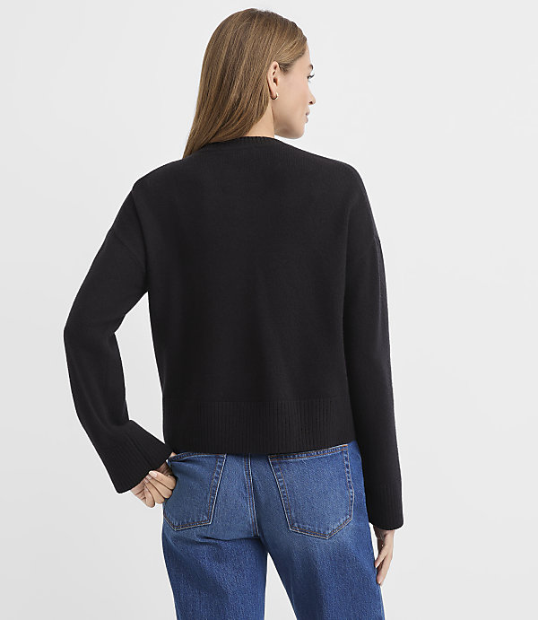 A woman wearing a black sweater and blue jeans stands with her back to the camera against a plain white background.