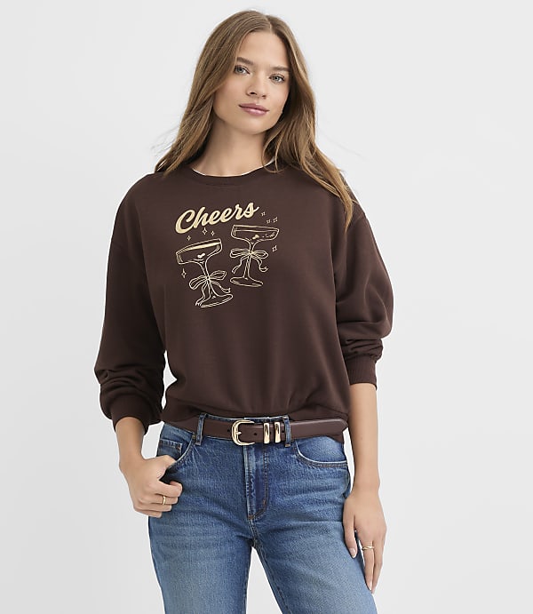 A woman wearing a brown "Cheers" sweatshirt and blue jeans stands against a plain white background.