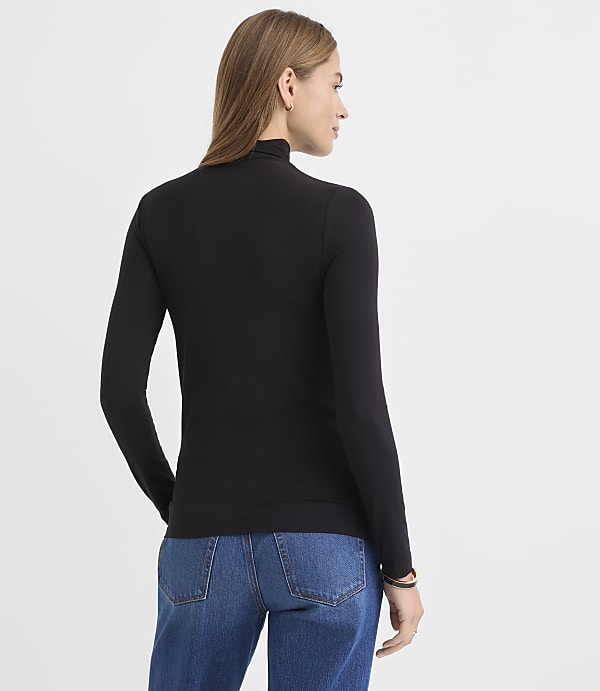 A woman wearing a black long-sleeved turtleneck and blue jeans stands with her back to the viewer against a plain white background.