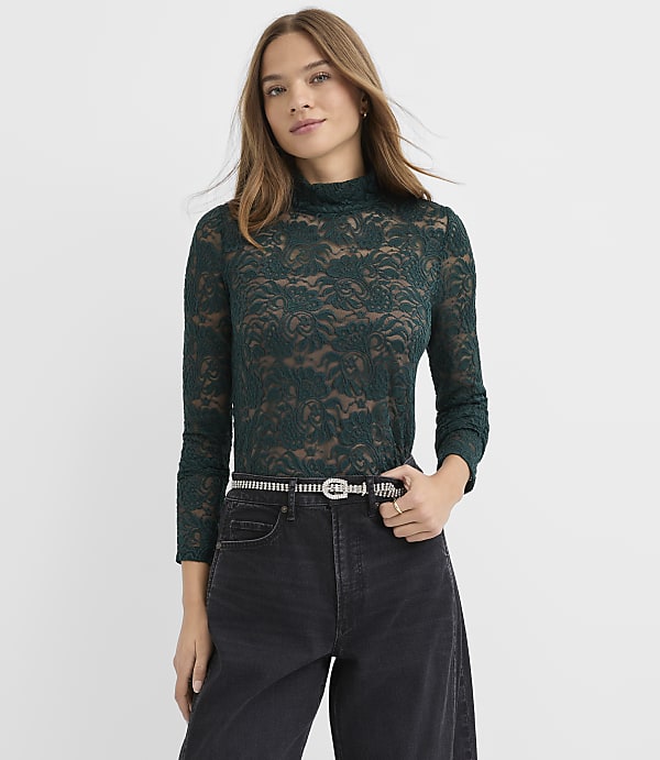 A woman wearing a dark green lace top and black jeans with a sparkly belt stands against a plain white background.