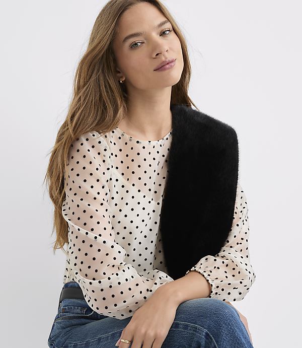 A woman wearing a polka-dot blouse and blue jeans poses with a black faux-fur stole against a plain white background.