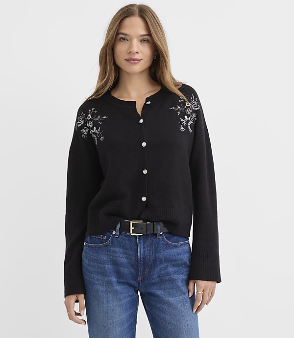 A woman wearing a black embroidered cardigan and blue jeans stands against a plain white background.