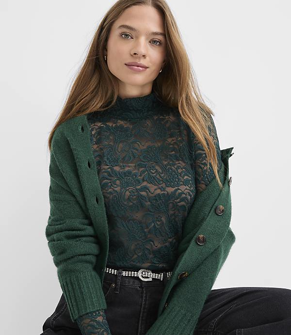 A woman wearing a dark green lace top and a matching green cardigan poses against a plain white background.