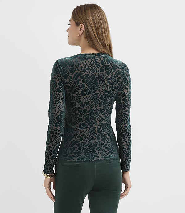 A woman wearing a green floral velvet top and matching pants stands against a plain white background.