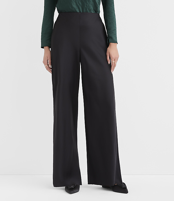 A person wearing a green top and wide-leg black trousers stands against a plain white background.