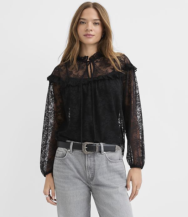 A woman wearing a black lace blouse and grey jeans stands against a plain white background.
