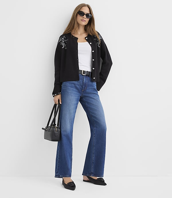 A woman wearing a black cardigan, white top, and flared blue jeans stands against a plain white background.