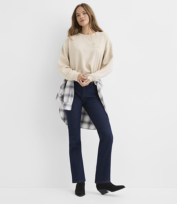 A woman wearing a beige sweatshirt, dark blue jeans, and black boots stands against a plain white background.