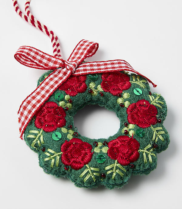A green fabric wreath adorned with red embroidered flowers and a red-and-white checkered bow rests on a plain white background.
