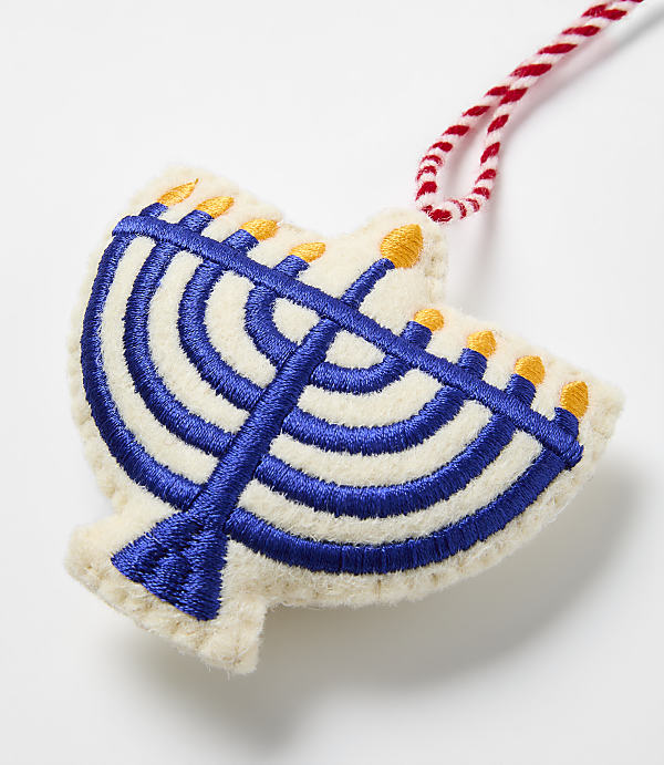 A blue embroidered menorah on a cream-colored felt ornament hangs against a plain white background.