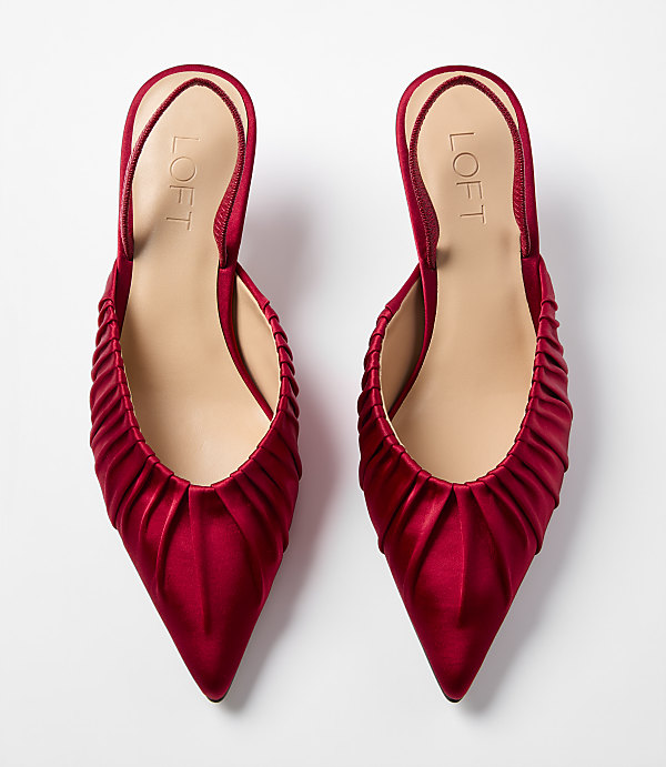 A pair of red satin pointed-toe mules with ruched detailing sits against a plain white background.