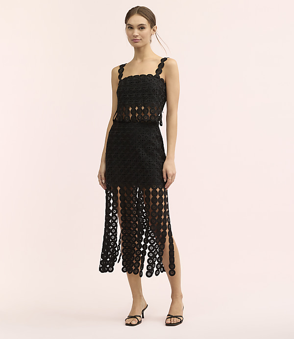 A woman wearing a black crochet dress stands against a plain, light pink background.