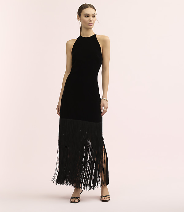 A woman wearing a black velvet halter dress with a long fringe skirt stands against a plain light pink background.