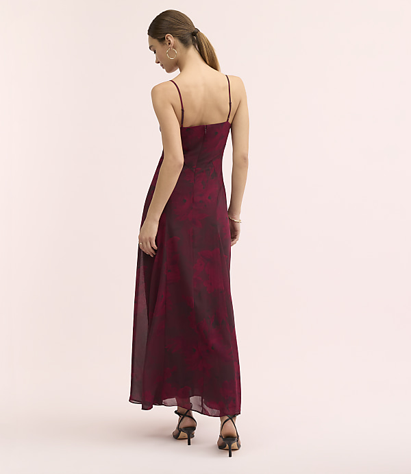 A woman in a long, floral burgundy dress and black heels stands against a plain, light pink background.