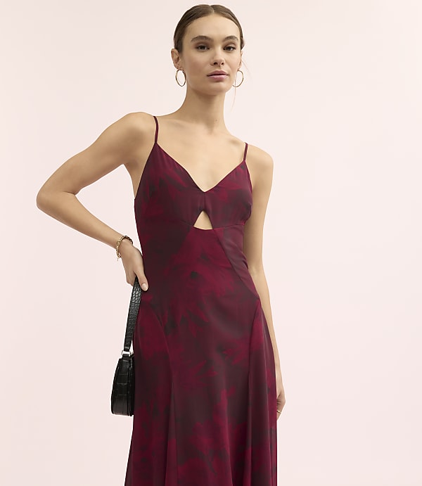 A woman wearing a maroon floral dress and holding a black handbag stands against a plain light pink background.