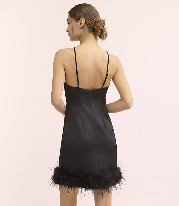 A woman wearing a black satin mini dress with feather trim stands with her back to the camera against a plain light pink background.