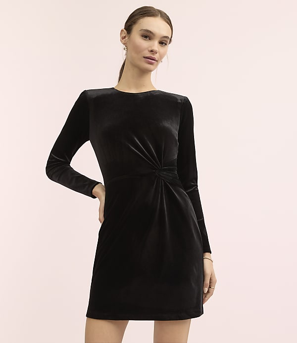 A woman wearing a long-sleeved black velvet dress stands against a plain light pink background.