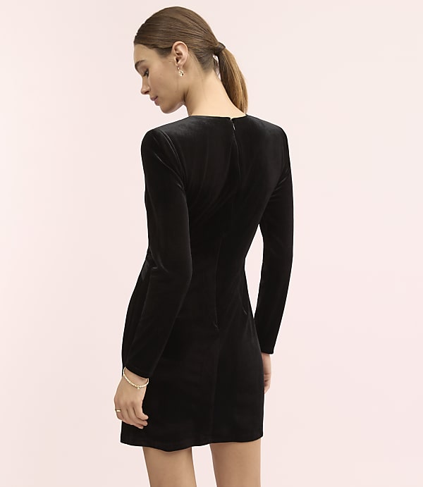 A woman wearing a black velvet long-sleeved mini dress stands against a plain light pink background.