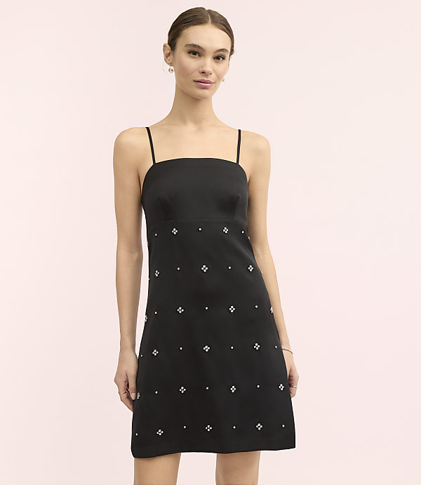 A woman wearing a black mini dress with small white embellishments stands against a plain light pink background.