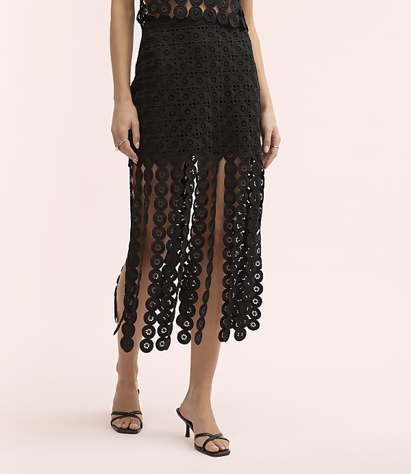 A person wears a black lace-patterned dress and black sandals against a plain light pink background.