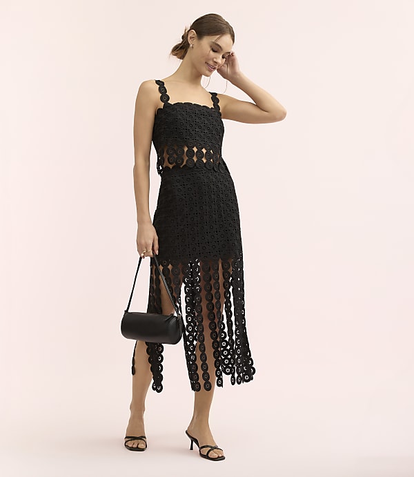 A woman wearing a black lace dress and holding a black handbag stands against a plain, light pink background.