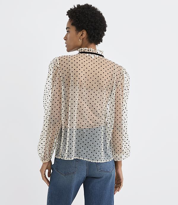 A woman wearing a sheer polka-dot blouse and blue jeans stands against a plain white background.