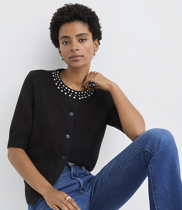 A woman wearing a black studded cardigan and blue jeans poses against a plain white background.